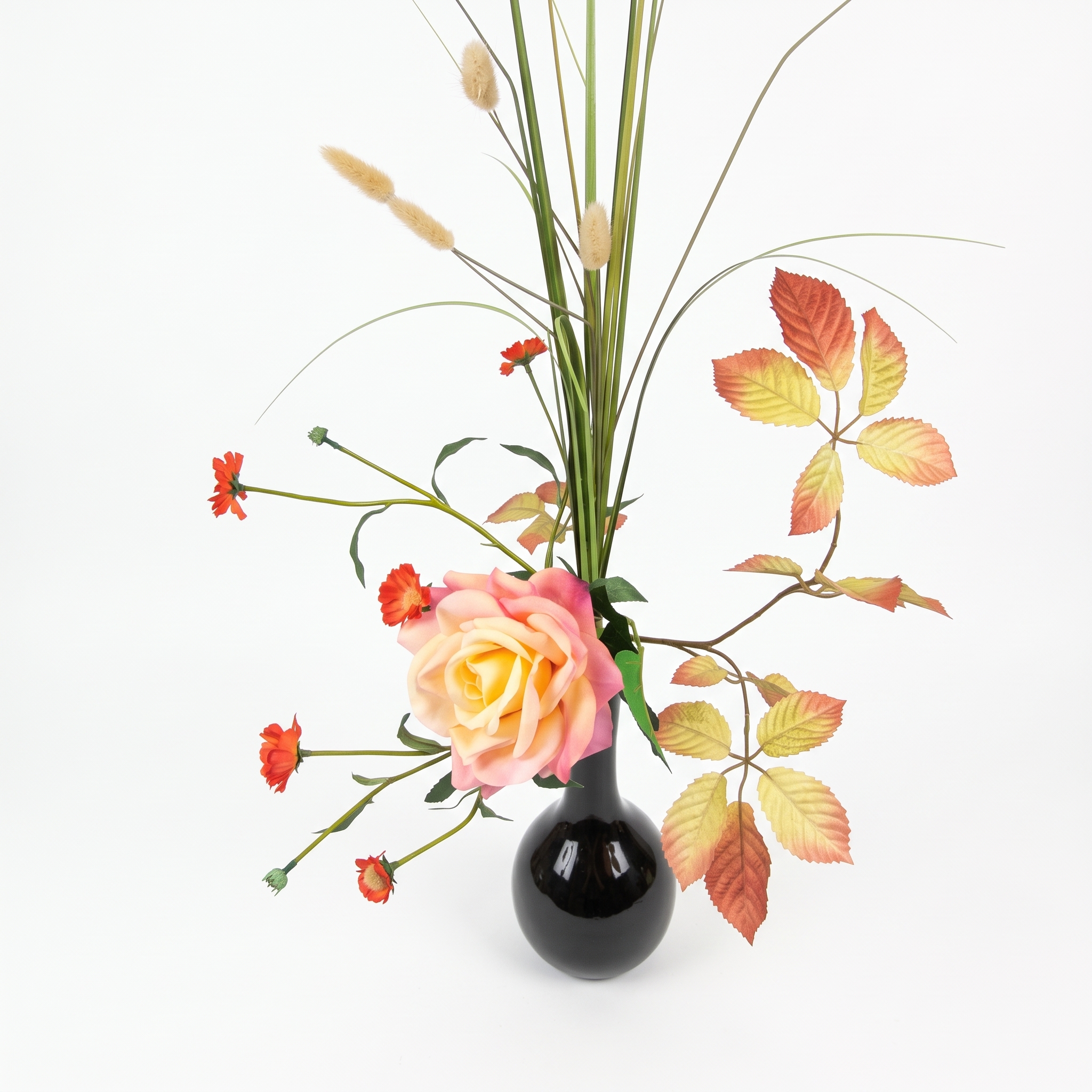 Bovenaanzicht van een asymmetrisch kunstbloemen arrangement met een geopende zijden roos, groene grassen en herfstkleurige bladtakken.