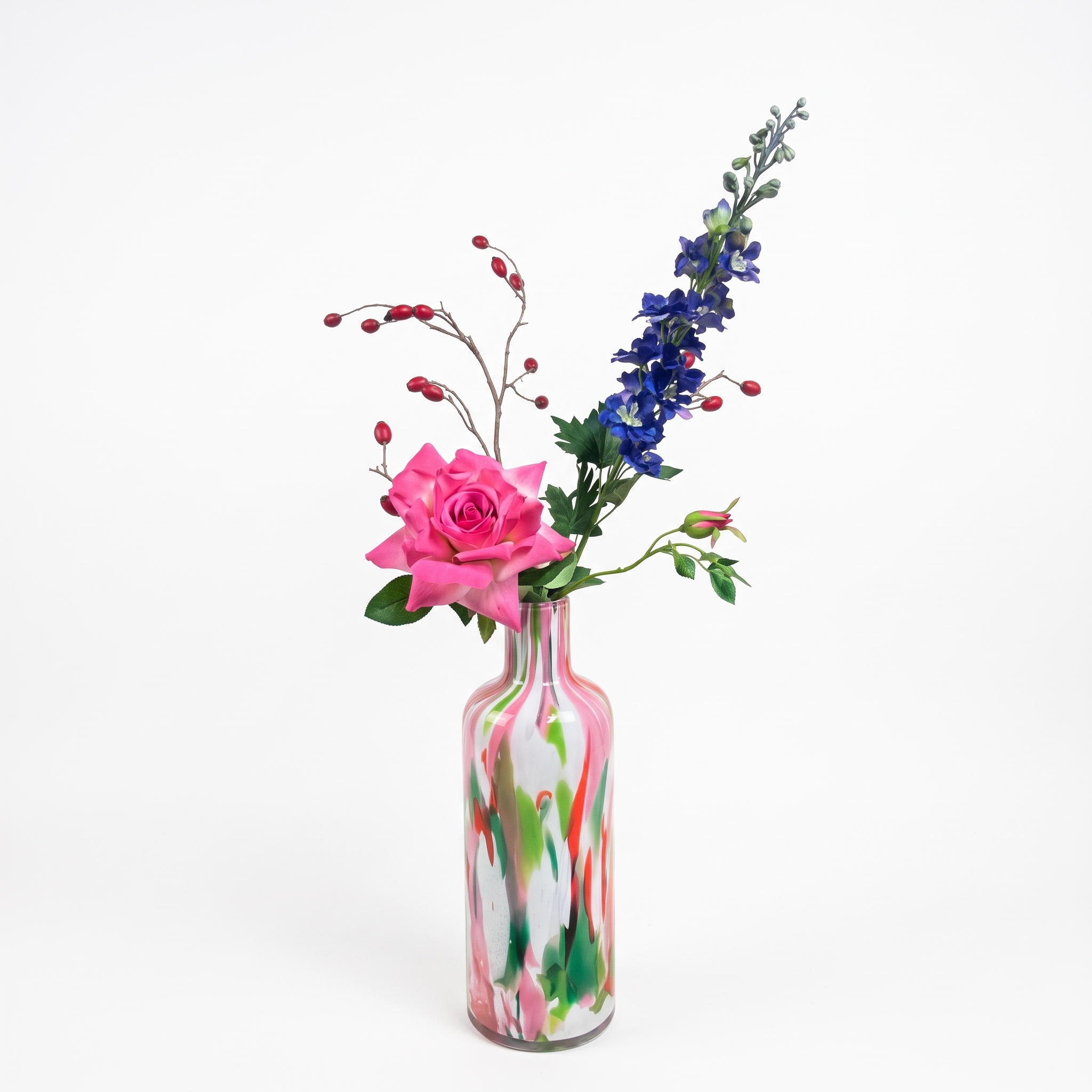 Een kunstboeket van een grote roze roos, blauwe ridderspoor en rode rozenbotteltak in een kleurrijke gemarmerde glazen vaas.