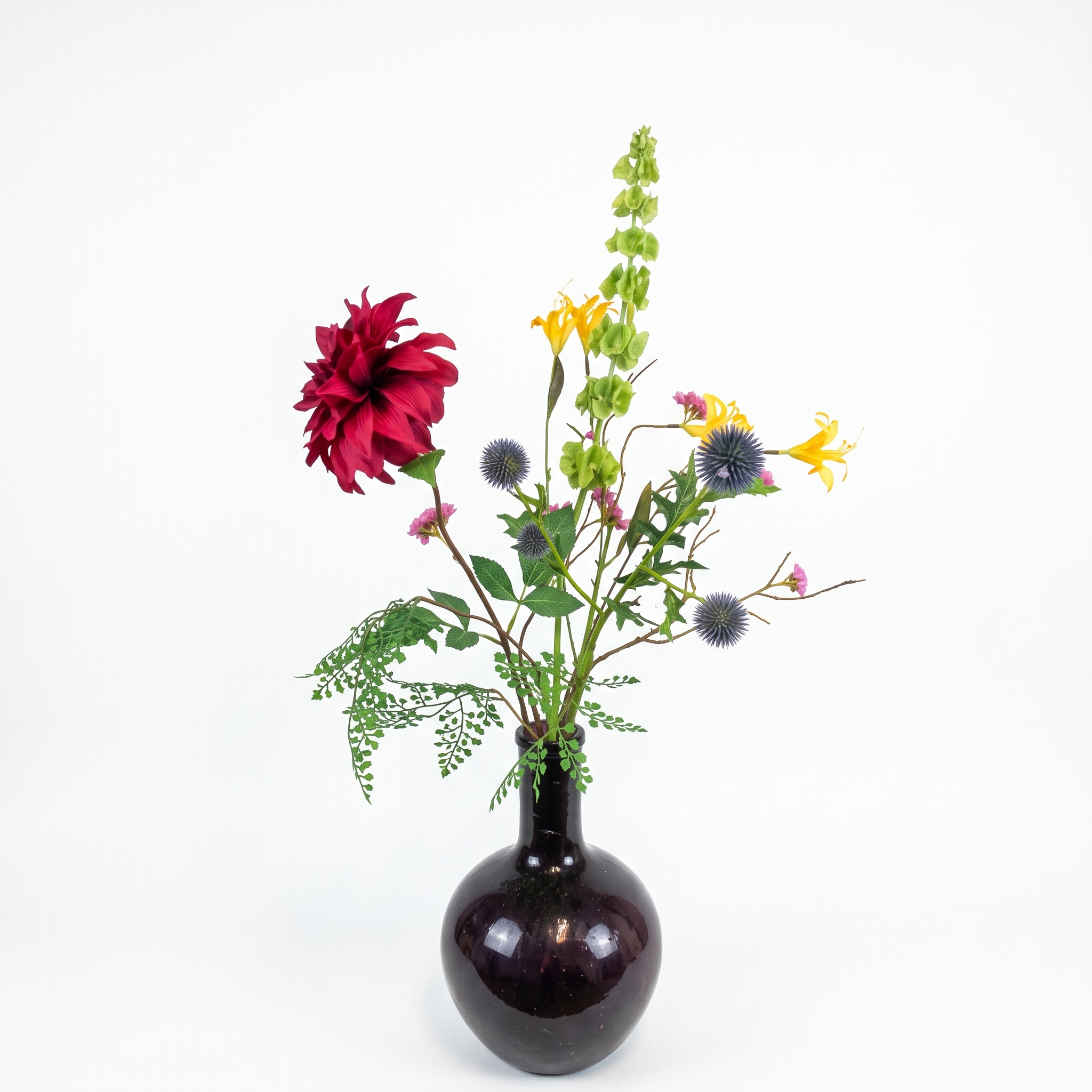 Kunstveldboeket met bordeauxrode dahlia, groene moluccella laevis en blauwe kogeldistel in een bolle donkere vaas